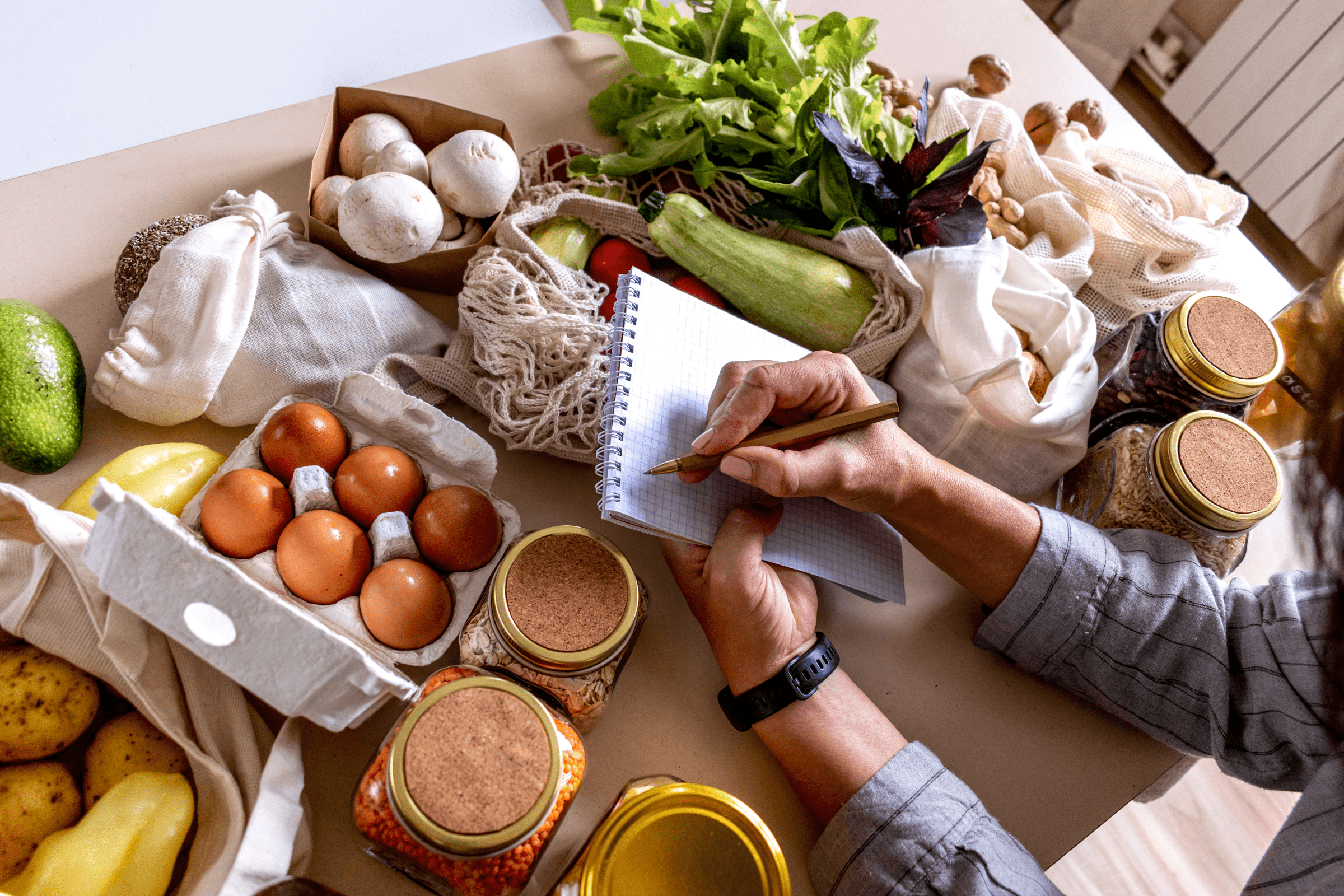 Person planlegger matinnkjøp hjemme med notatblokk og matvarer på bordet.