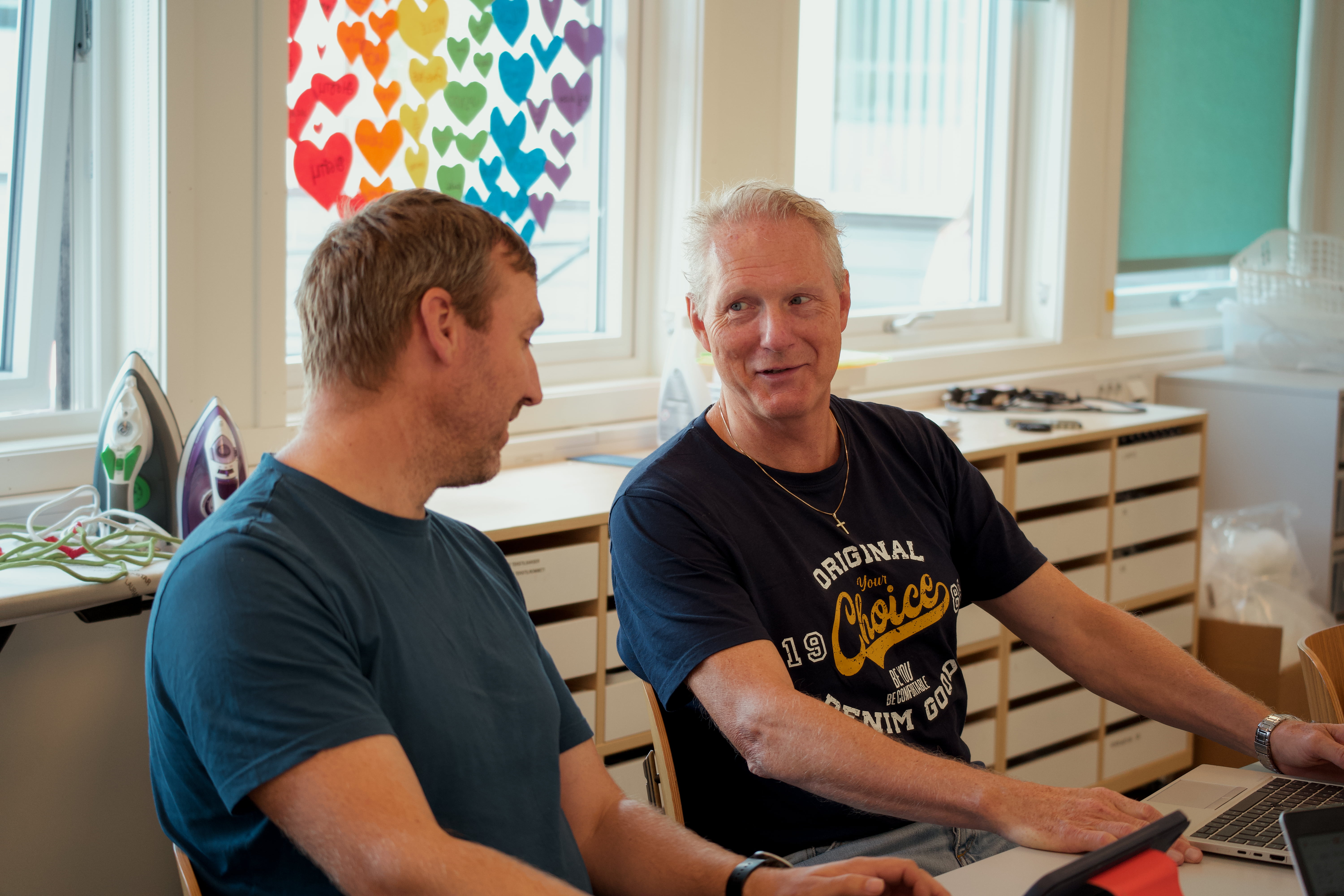 To personer sitter ved et bord og jobber på en bærbar PC i et rom med store vinduer. På veggen henger en plakat med fargerike hjerter, og det står strykejern på en hylle bak dem.